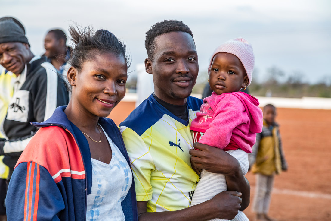 In this image, Bito Billa is standing next to his wife. He is holding his baby daughter (two years old) in his arms, and his wife has an expression of pride and joy. Corumana FC in their yellow kit for the 2019 Rhino Cup Champions League season won against Chelsea FC on the Corumana soccer field, Mozambique. The Corumana FC team is sponsored by Wild and Free Foundation.