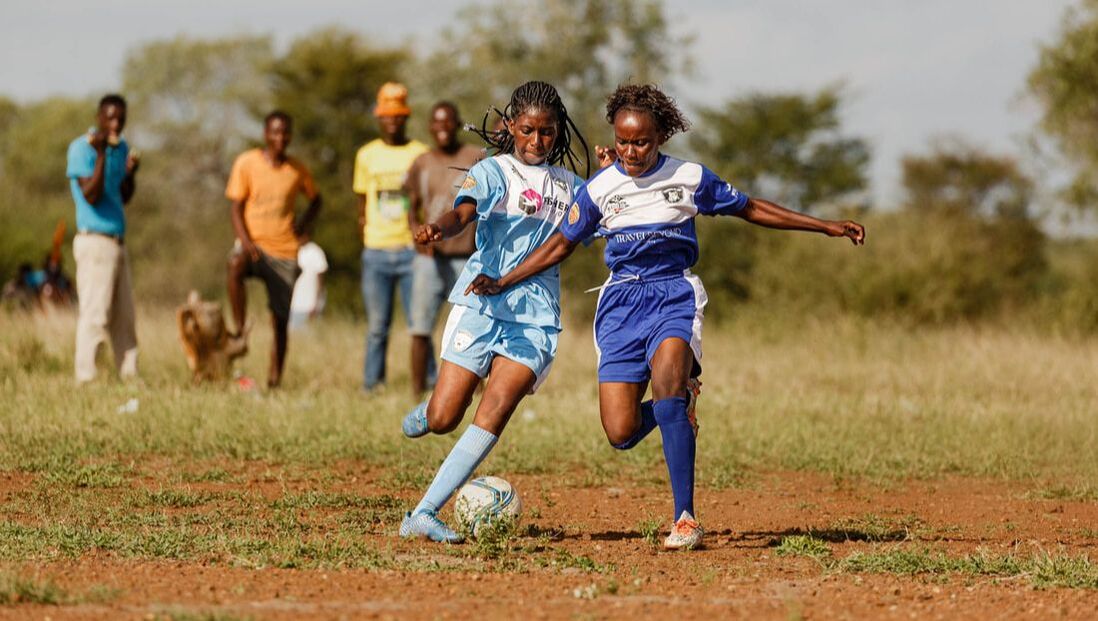Two women football players from the RCCL Mozambique Girl’s League engaged in a competitive match during the beginning of the 2022 season. The light blue football team is called the Mulheres de Baptine Buffalos, they are sponsored by Fisher Futbol, a private family group from the USA. The football team known as the Perigrinas de Sabié Lions is distinguished by its darker blue color. The Lions have been proudly sponsored by Travel Beyond, an international travel agency, since 2019.